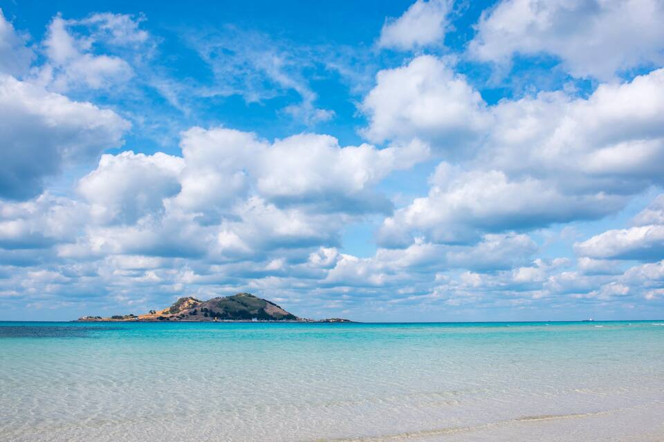 Une plage à l’eau turquoise et limpide, un rivage sablonneux et, au loin, une île surmontée d’une colline.