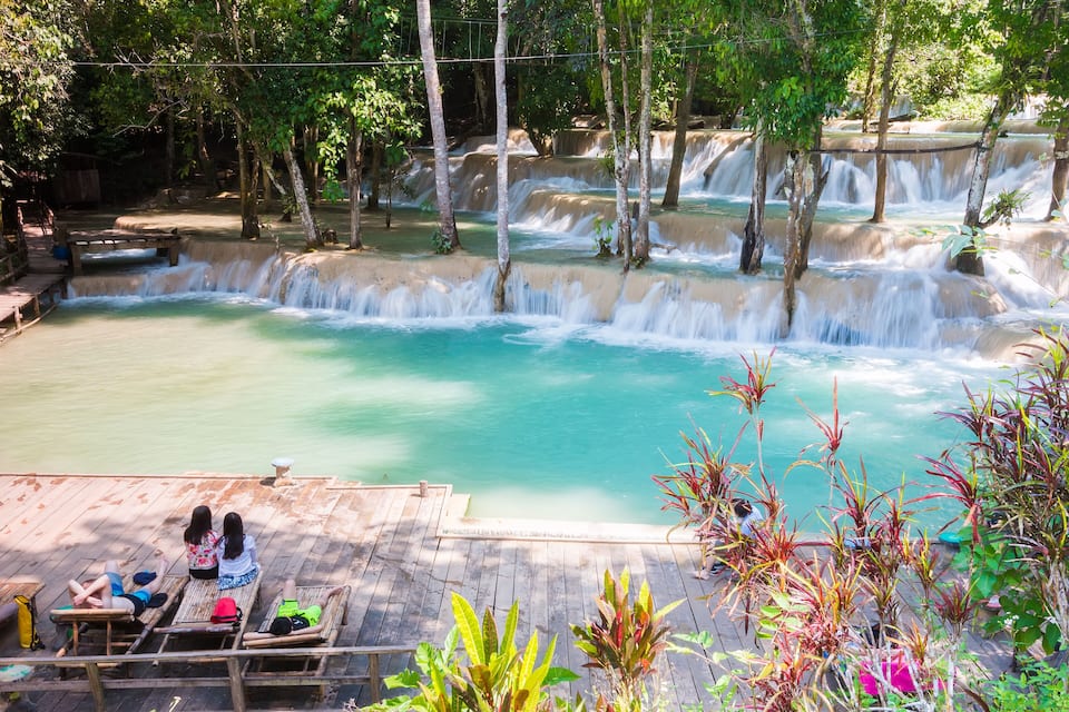 A waterfall with multiple tiers, surrounded by lush greenery and a wooden deck with people relaxing.