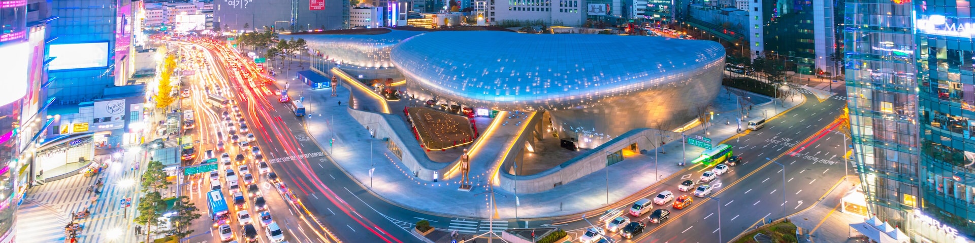 A modern urban intersection with a distinctive curved building, busy traffic, and illuminated billboards.