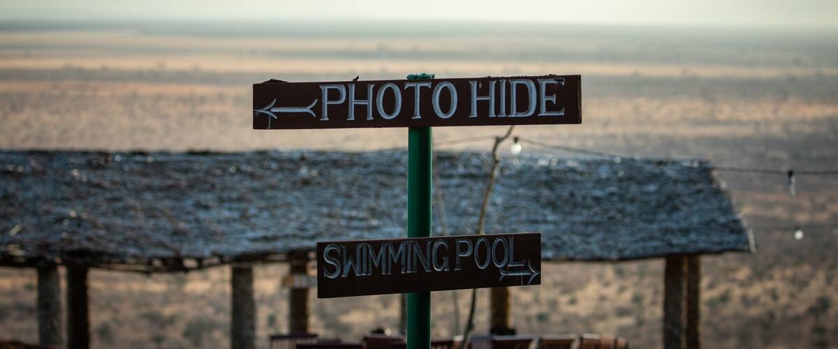 A signpost with 'Photo Hide' and 'Swimming Pool' directions, overlooking a beach with lounge chairs.