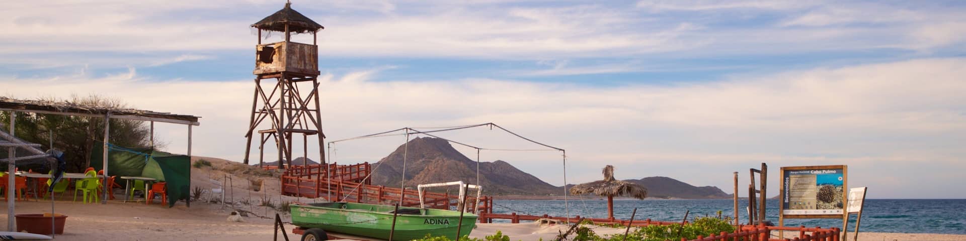 Una playa con una torre de observación de madera, un bote verde, una pasarela de madera roja y un cartel.