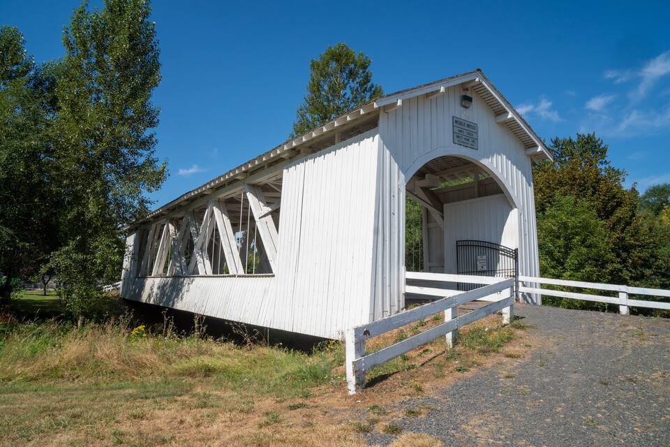 Eine weiße gedeckte Brücke mit Metallgeländer, umgeben von Bäumen und Gras.