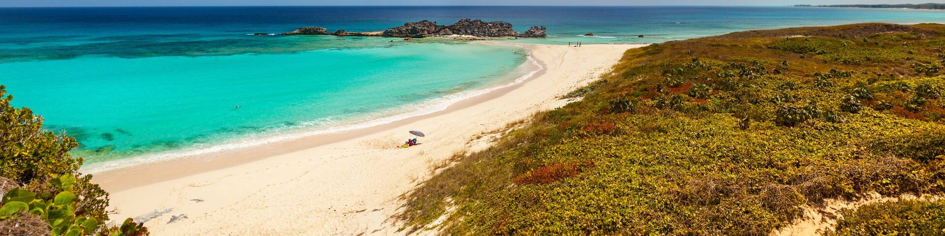 Una playa con aguas turquesas cristalinas, arena blanca y vegetación verde.