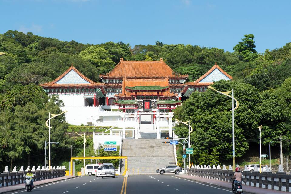 A traditional Chinese gate with ornate architecture, a road in front, and lush green trees in the background.