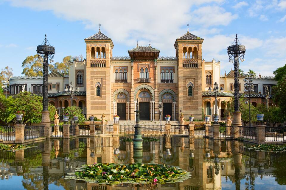 Um edifício histórico com arquitetura ornamentada, um lago espelhado com folhas de nenúfar e um céu claro.
