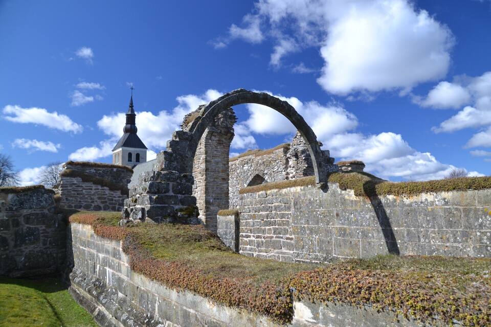An ancient stone wall with a large archway and a church with a spire in the background.