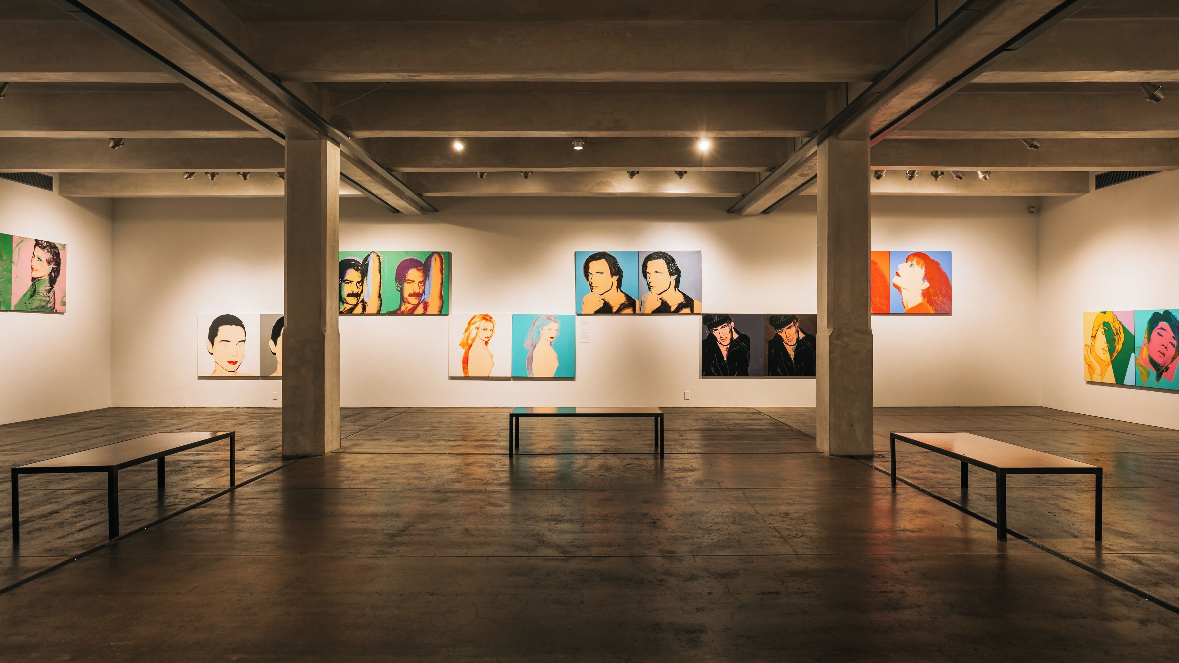 Een kunstgalerie met schilderijen aan de muur en banken in het midden.