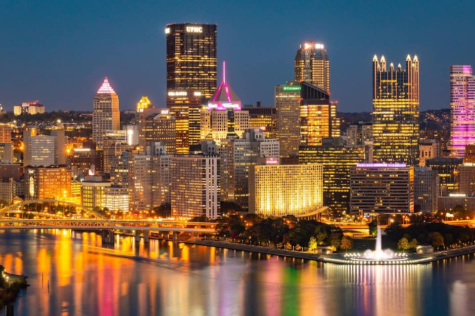 A city skyline at night with illuminated buildings and a river reflecting the lights.