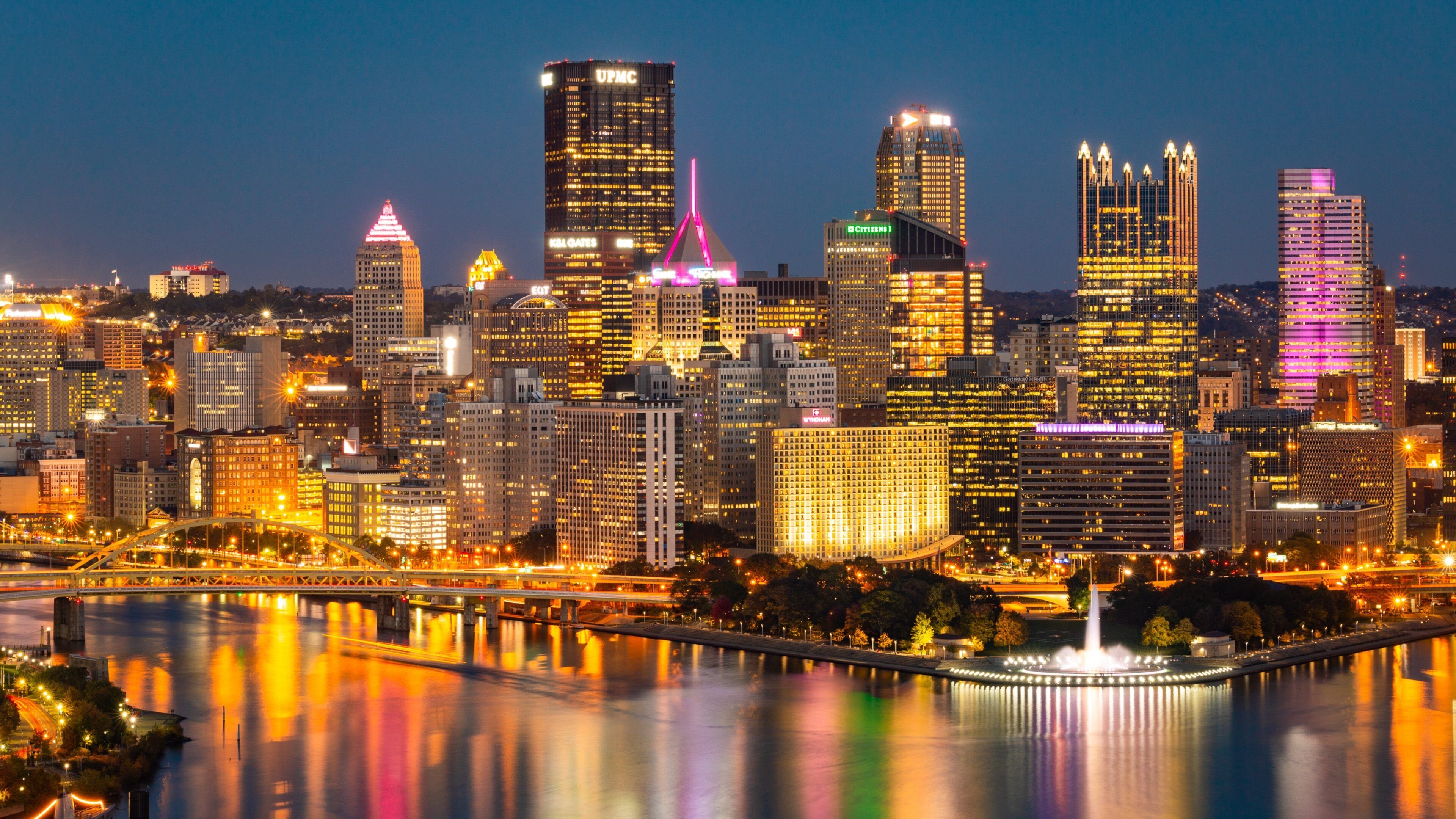 A city skyline at night with illuminated buildings and a river reflecting the lights.
