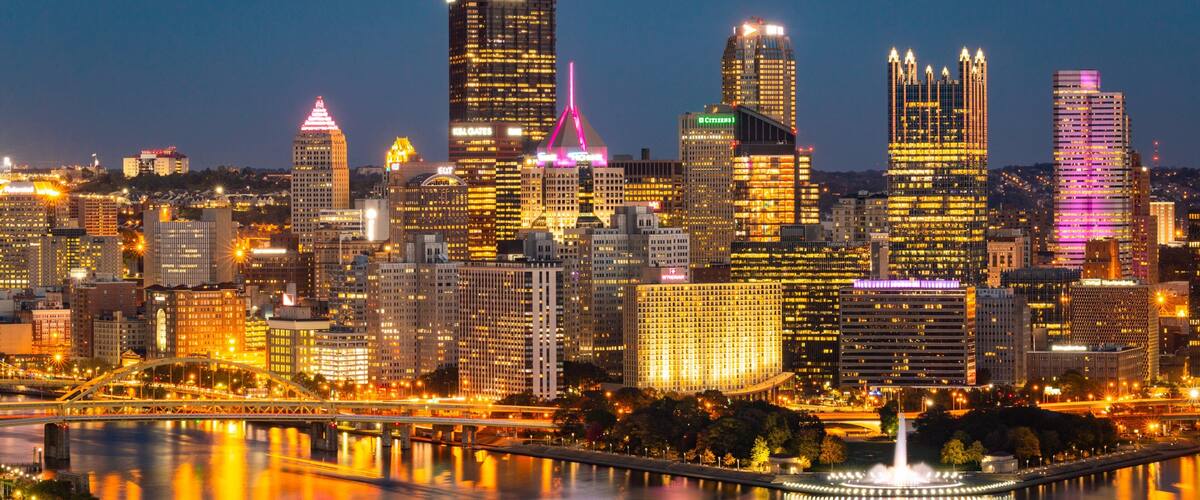 A city skyline at night with illuminated buildings and a river reflecting the lights.