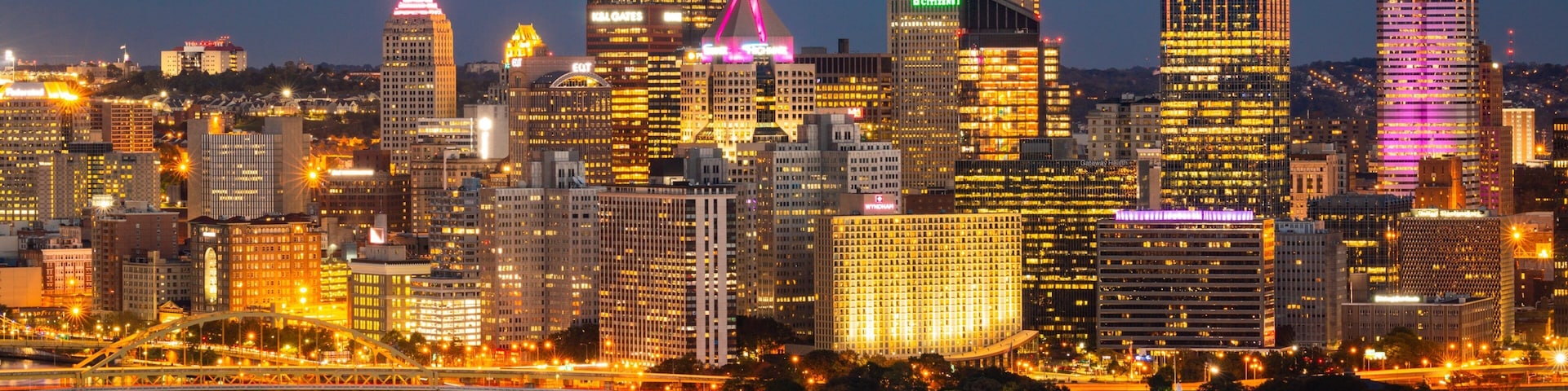 A city skyline at night with illuminated buildings and a river reflecting the lights.