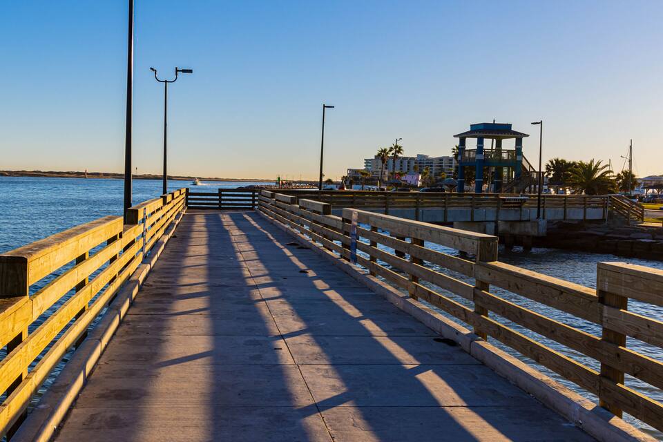A wooden pier extending into calm waters, with a lighthouse in the distance and a blue building on the right.