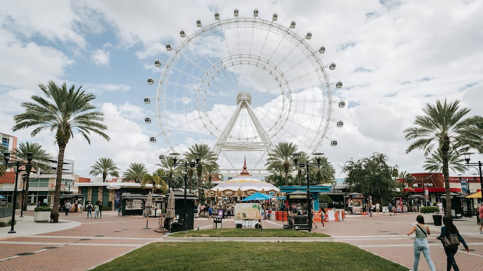 A large Ferris wheel, palm trees, and a plaza with shops and people.