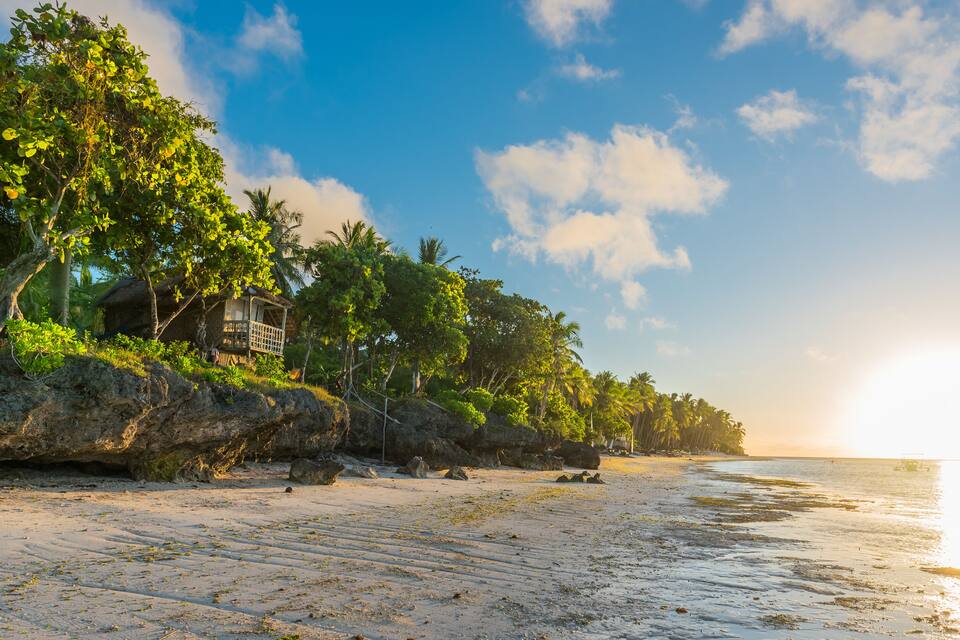 Una playa con una casa de madera sobre un promontorio rocoso, rodeada de palmeras y olas del océano.