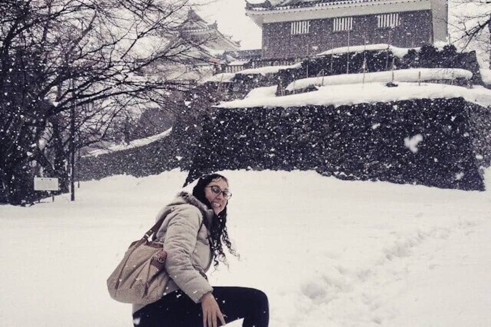 A person crouching in the snow with a traditional Japanese building in the background.