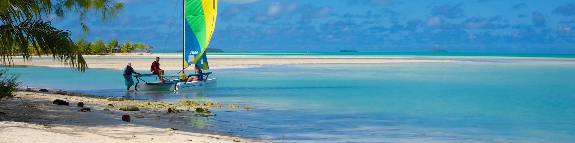 A sailboat with a colorful sail on a beach with clear turquoise water and palm trees.