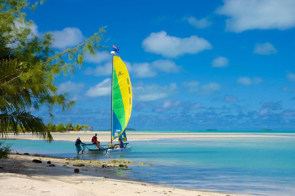 A sailboat with a colorful sail on a beach with clear turquoise water and palm trees.