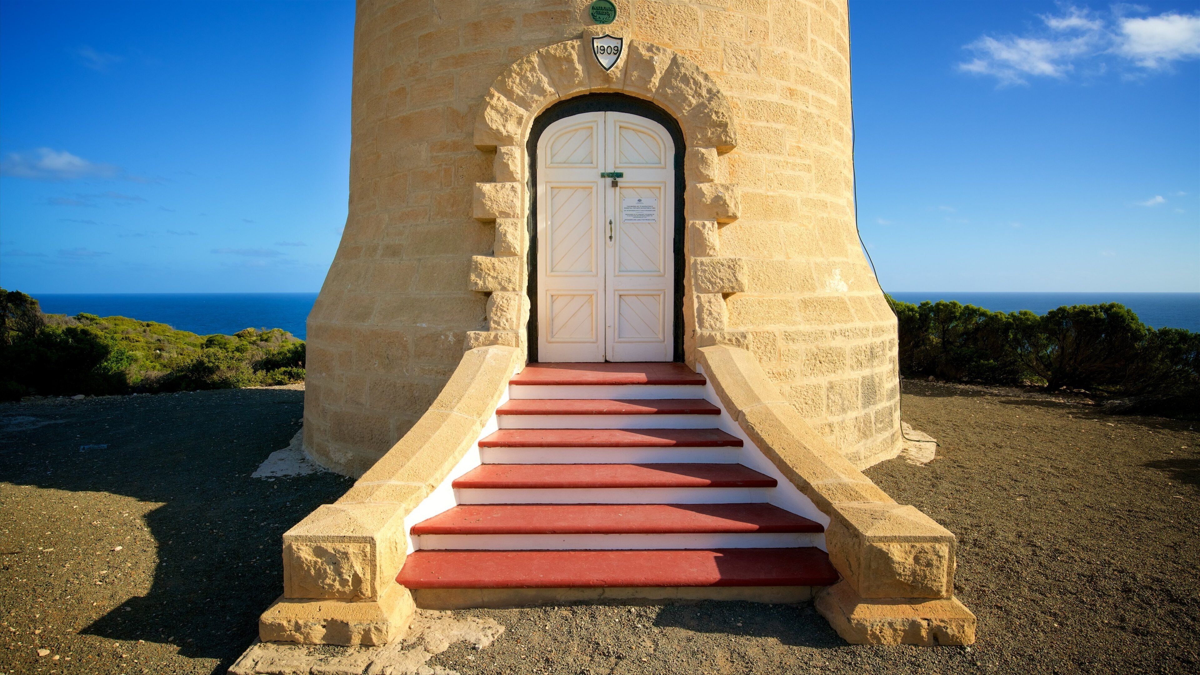 A stone tower with a white door and red steps leading up to it, overlooking a coastal landscape.