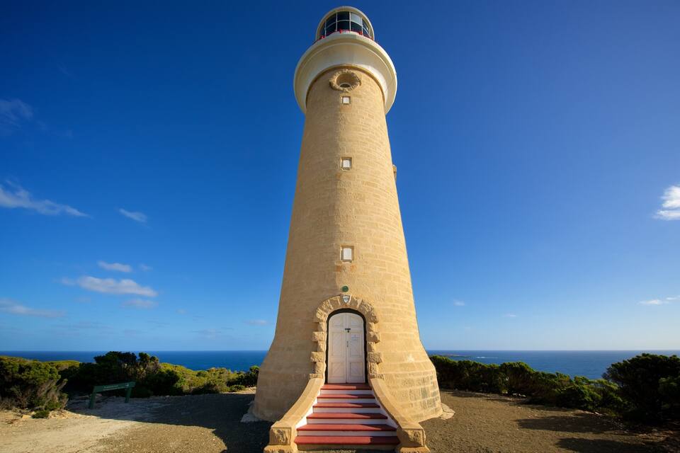 A tall, beige lighthouse with a red staircase leading to a white door, surrounded by greenery and the ocean under a clear blue sky.