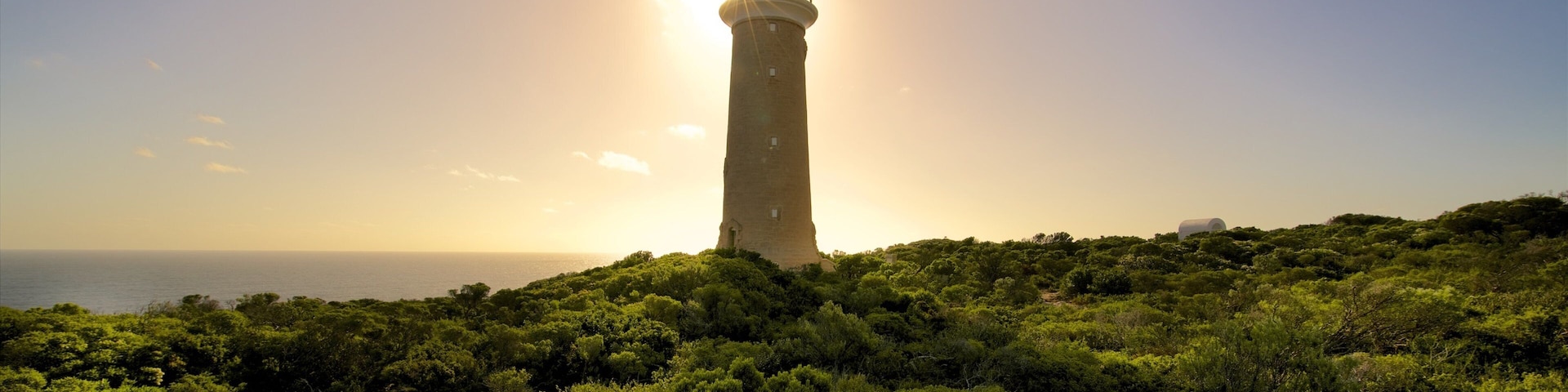 A lighthouse on a hill with dense greenery and the sun setting in the background.