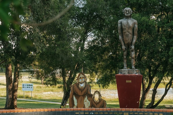 Eine Statue eines Mannes und eines Kindes in einem Park mit Bäumen und einem Schild im Hintergrund.