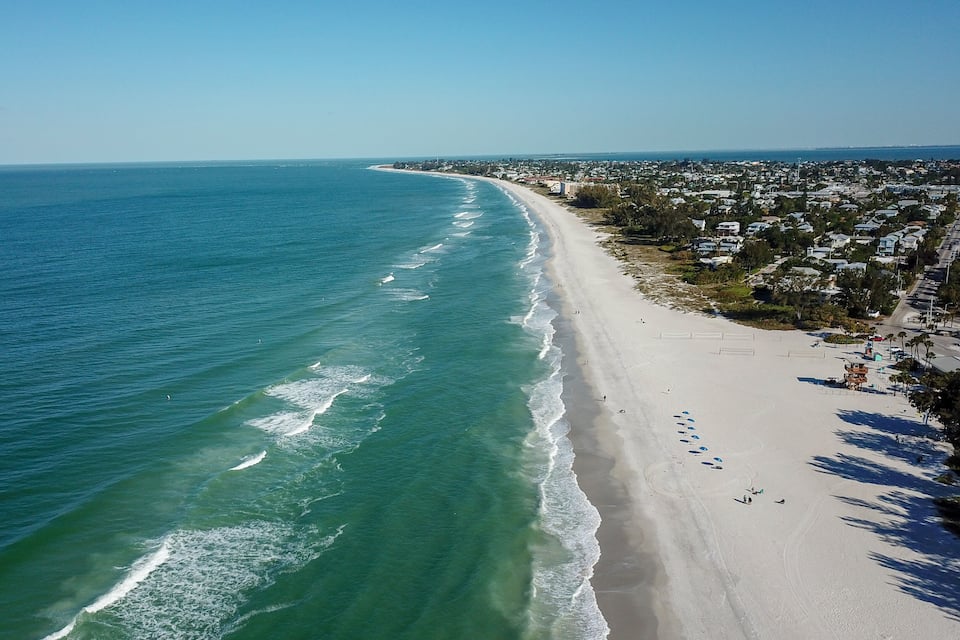 A coastal town with a long sandy beach, clear blue ocean, and residential buildings along the shore.