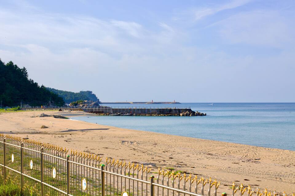 A sandy beach with a wooden fence, calm water, and a rocky outcrop in the distance.