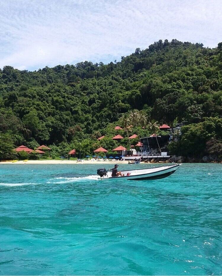 A boat on turquoise water with a beach and red-roofed structures in the background.