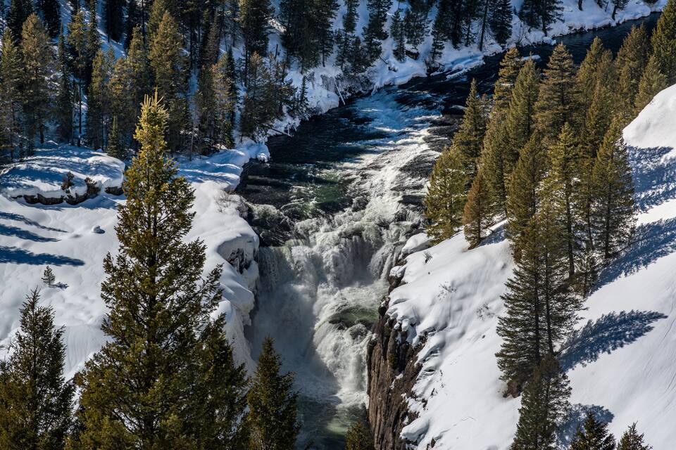A snow-covered river canyon with a waterfall surrounded by evergreen trees.