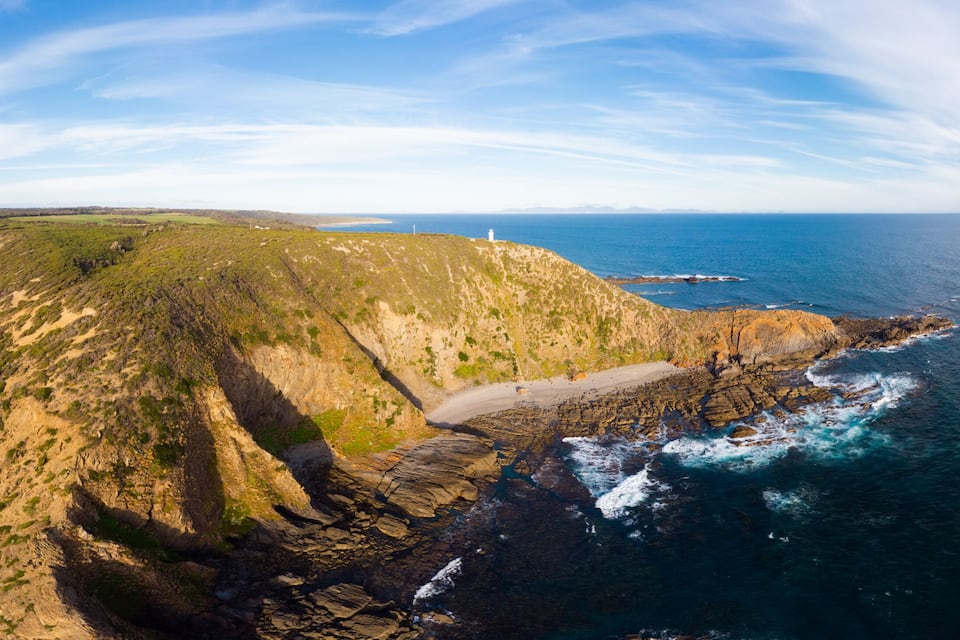 Paesaggio costiero con un faro, scogliere rocciose e una spiaggia.
