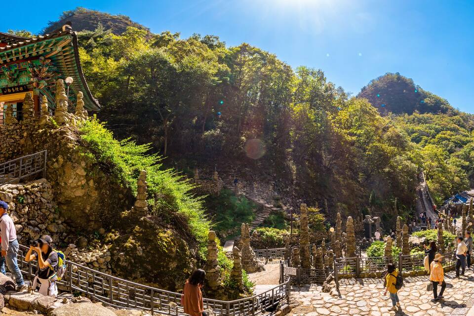 A temple complex with traditional architecture, stone statues, and a mountainous backdrop.