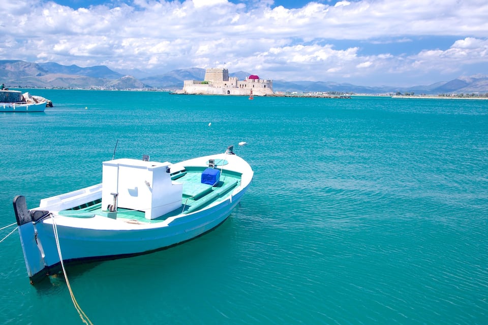 Une mer turquoise, avec un petit bateau amarré près d’un phare et, au loin, un château historique.