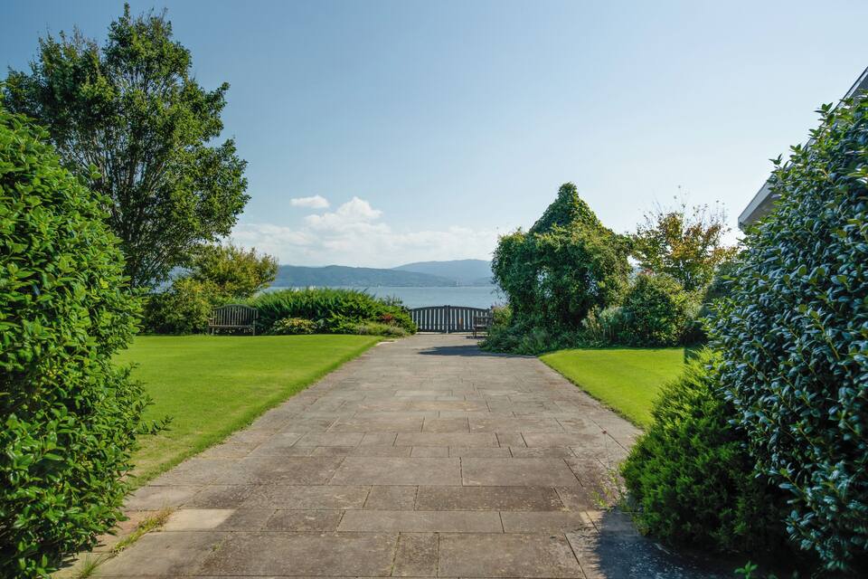 A paved pathway leading to a gazebo in a well-maintained garden with lush greenery and a view of distant hills.