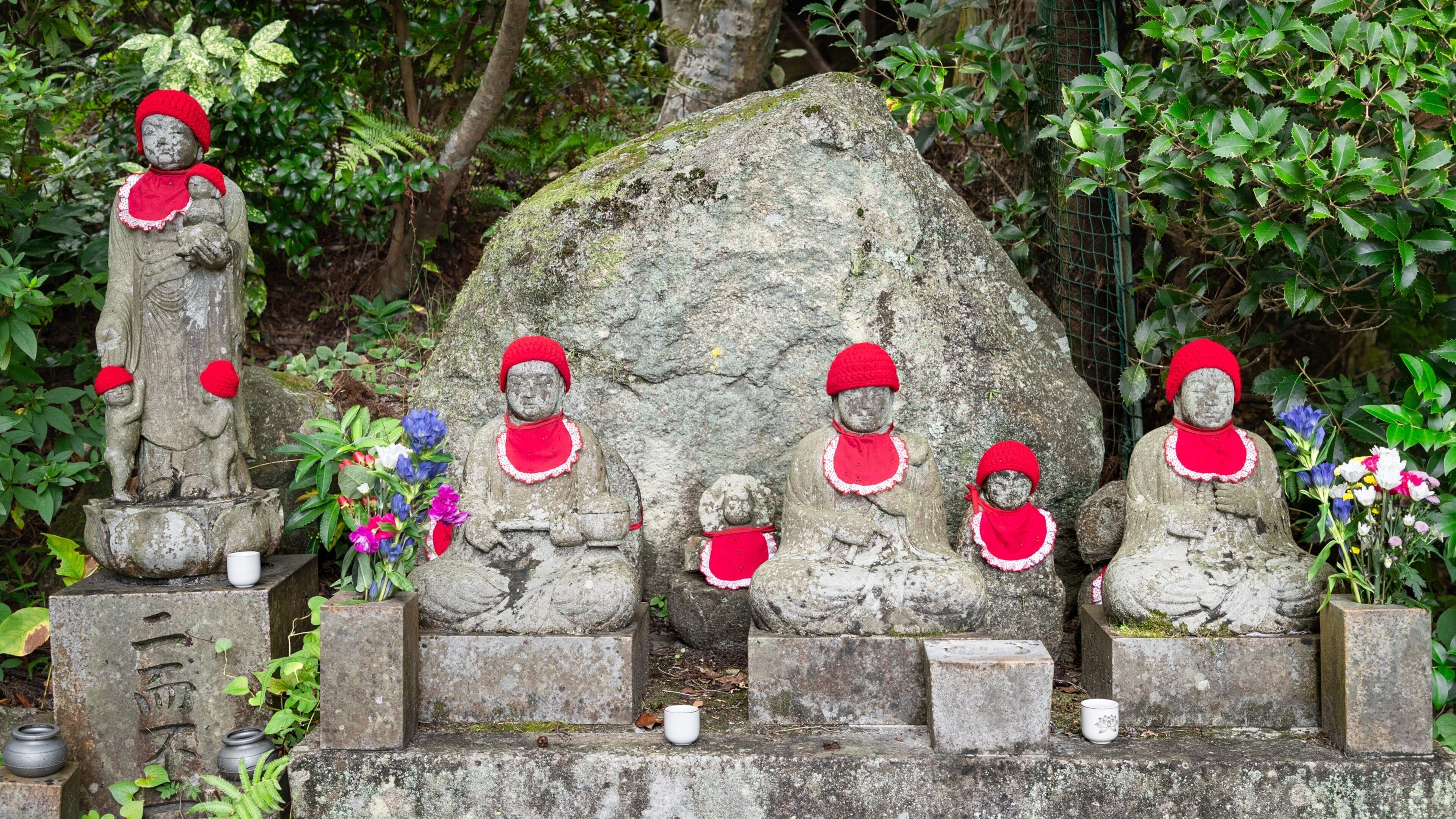Stone statues of Buddha in a garden, surrounded by greenery and flowers.