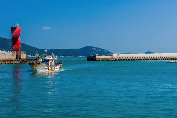 Fishing boat sailing near a vibrant red lighthouse in a harbor.