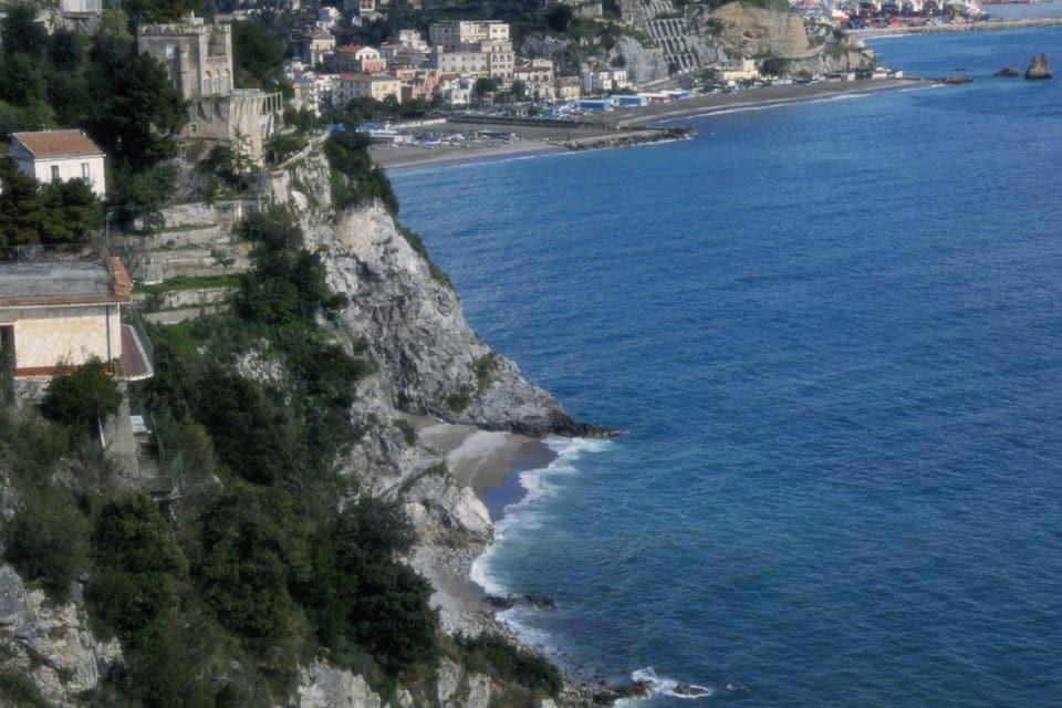 A coastal town with buildings on a cliff overlooking the sea, a bridge in the distance, and mountains in the background.