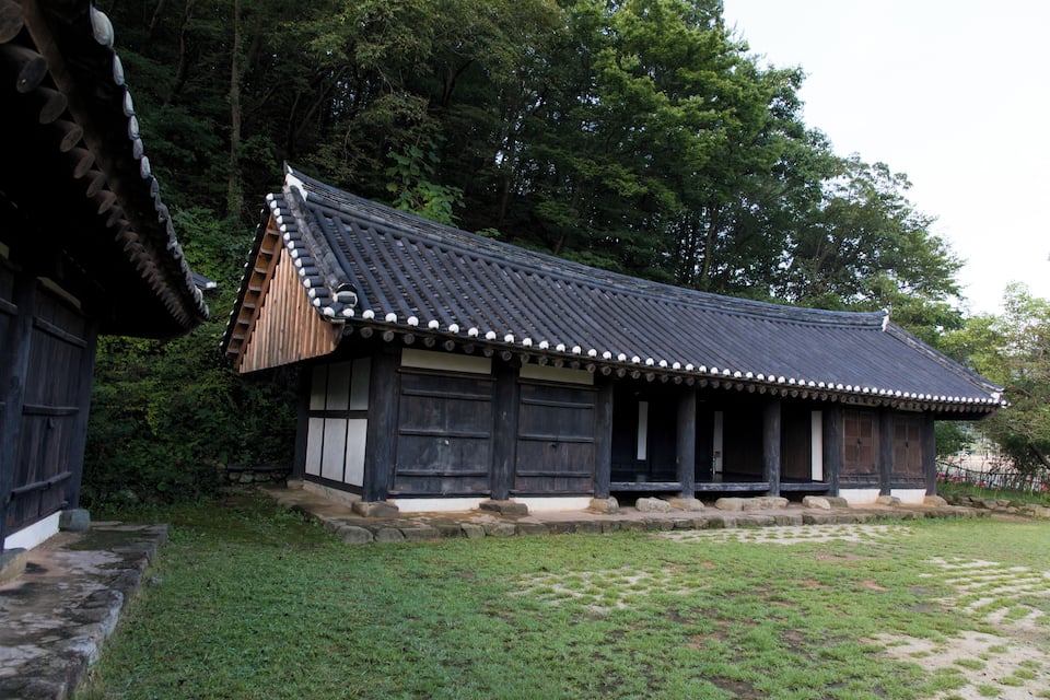 A traditional Korean building with a tiled roof and wooden walls, surrounded by a grassy area and trees.