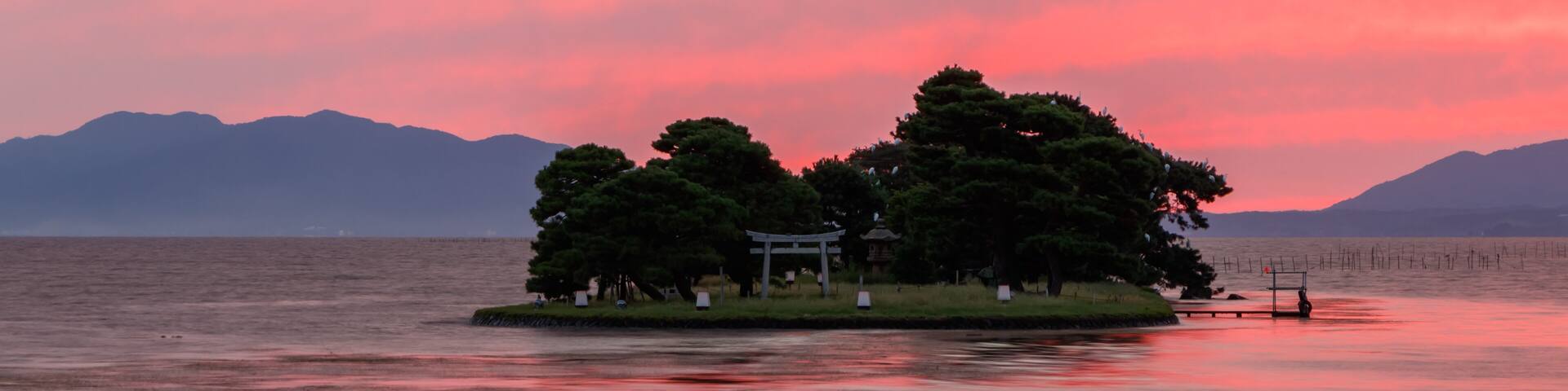 A tranquil lake at sunset with a small island featuring a gazebo and a torii gate.