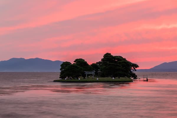 A tranquil lake at sunset with a small island featuring a gazebo and a torii gate.