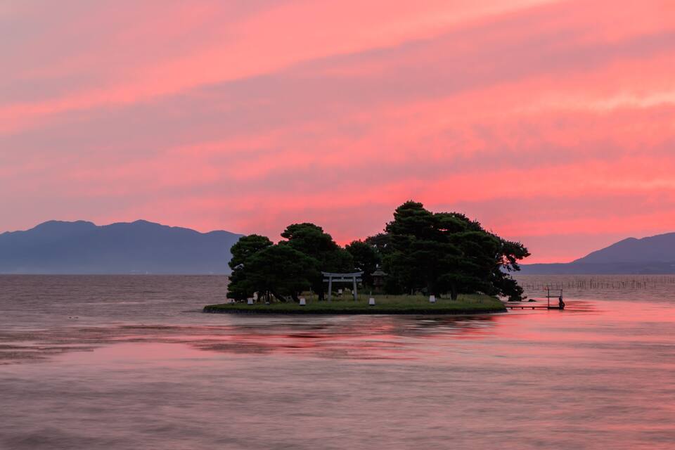 Un lago tranquilo al atardecer con una pequeña isla que tiene un pabellón y un torii.