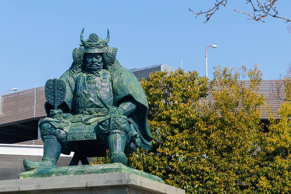 A statue of a warrior seated on a pedestal, with a sign in the background and trees in the foreground.