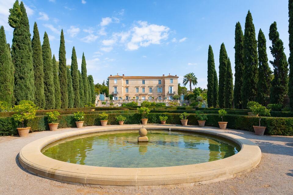 A formal garden with a circular pond, tall cypress trees, and a classical building in the background.