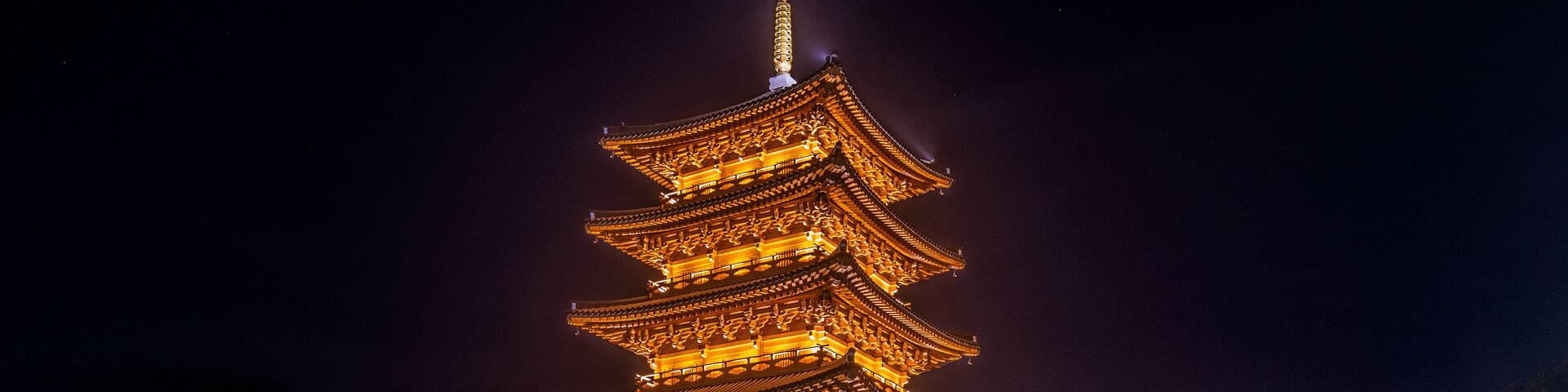A traditional pagoda with multiple tiers, illuminated at night, surrounded by a stone wall and a small bridge.