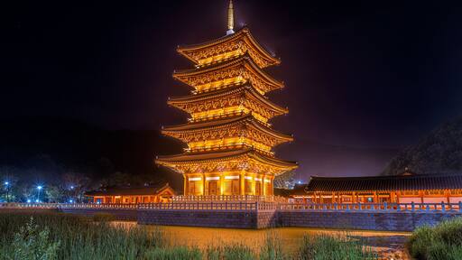 A traditional pagoda with multiple tiers, illuminated at night, surrounded by a stone wall and a small bridge.