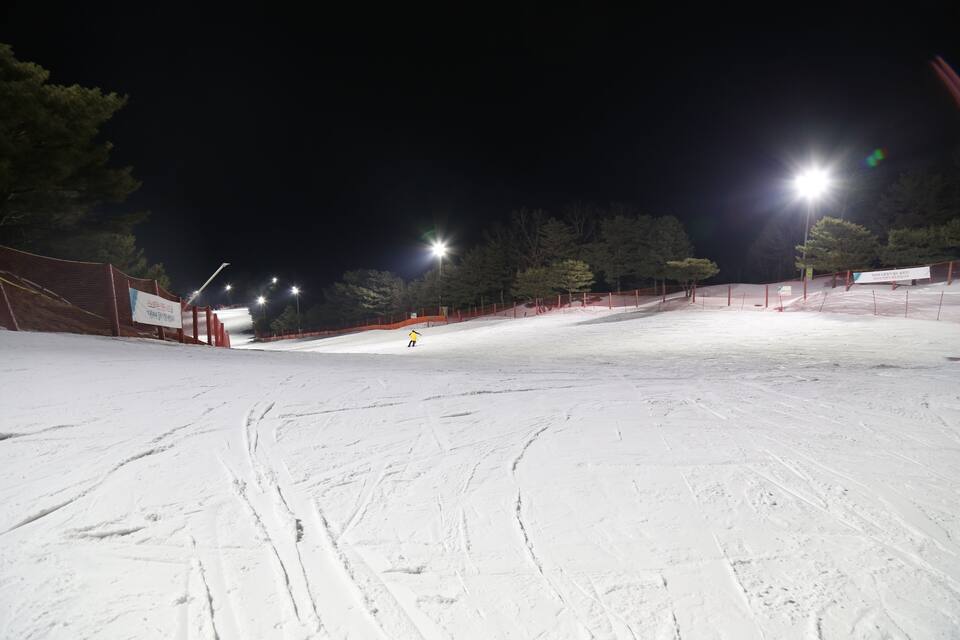 A snow-covered ski slope at night with artificial lighting and orange safety barriers.