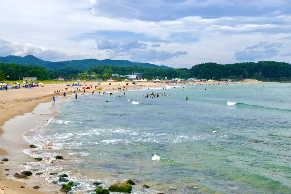Uma praia com águas cristalinas, pedras e montanhas ao fundo.