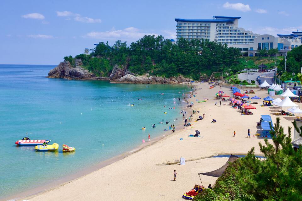 A beach with clear turquoise water, a sandy shore, and a forested hill in the background.