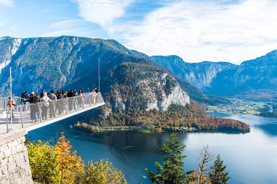 A scenic overlook with a group of people on a bridge, a lake, and surrounding mountains.