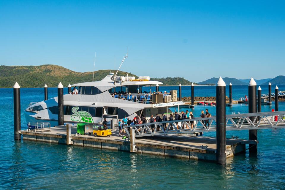 A large yacht docked at a pier with people on board and on the pier.