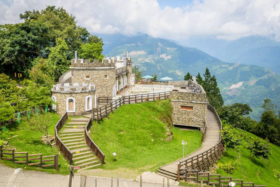 A stone castle with a wooden spiral staircase, surrounded by lush greenery and mountains in the background.